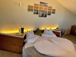 Um quarto com cama, mesas de cabeceira, luminária na parede e uma série de quadros retratando o pôr do sol sobre o mar.