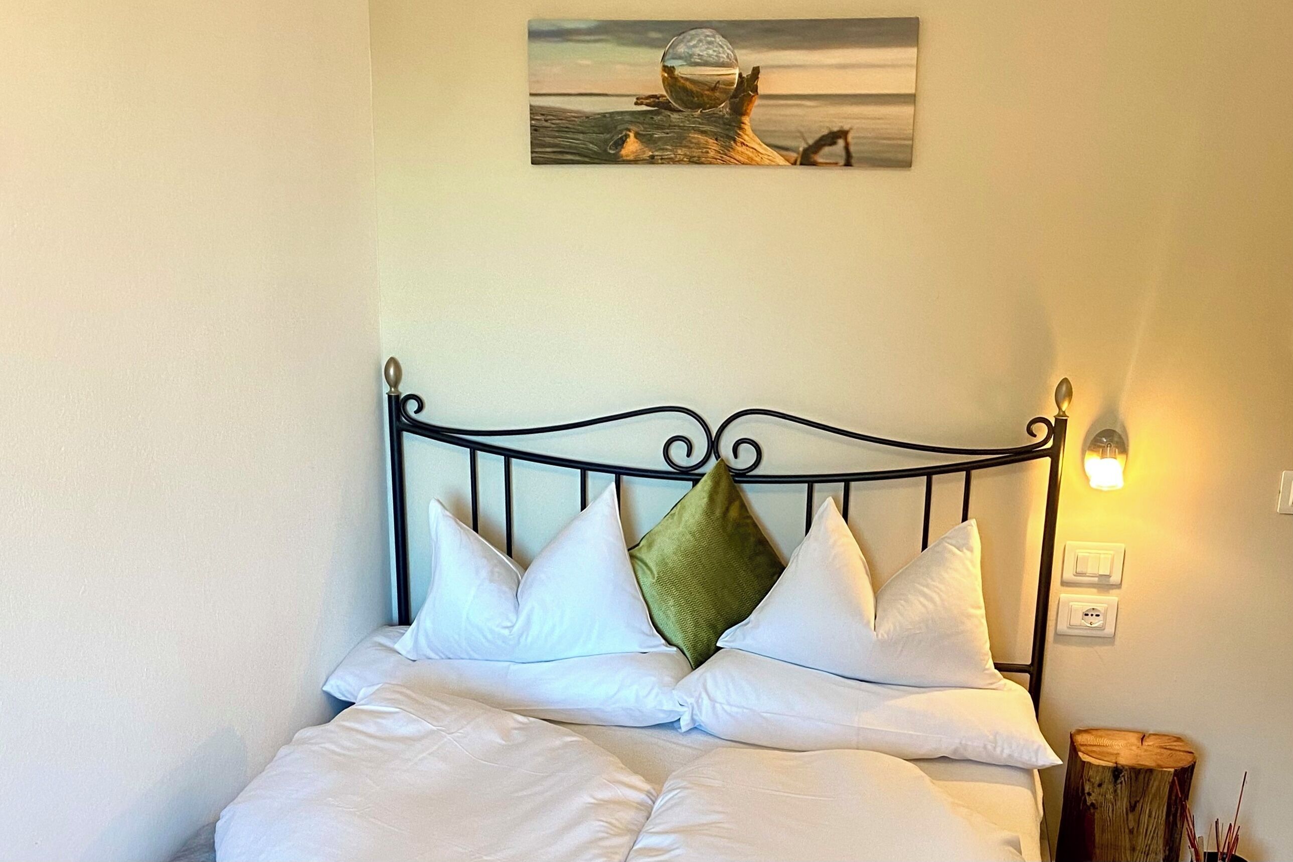 A neatly made bed with white bedding and a single green pillow, a wall-mounted picture of a beach scene, and a bedside lamp.