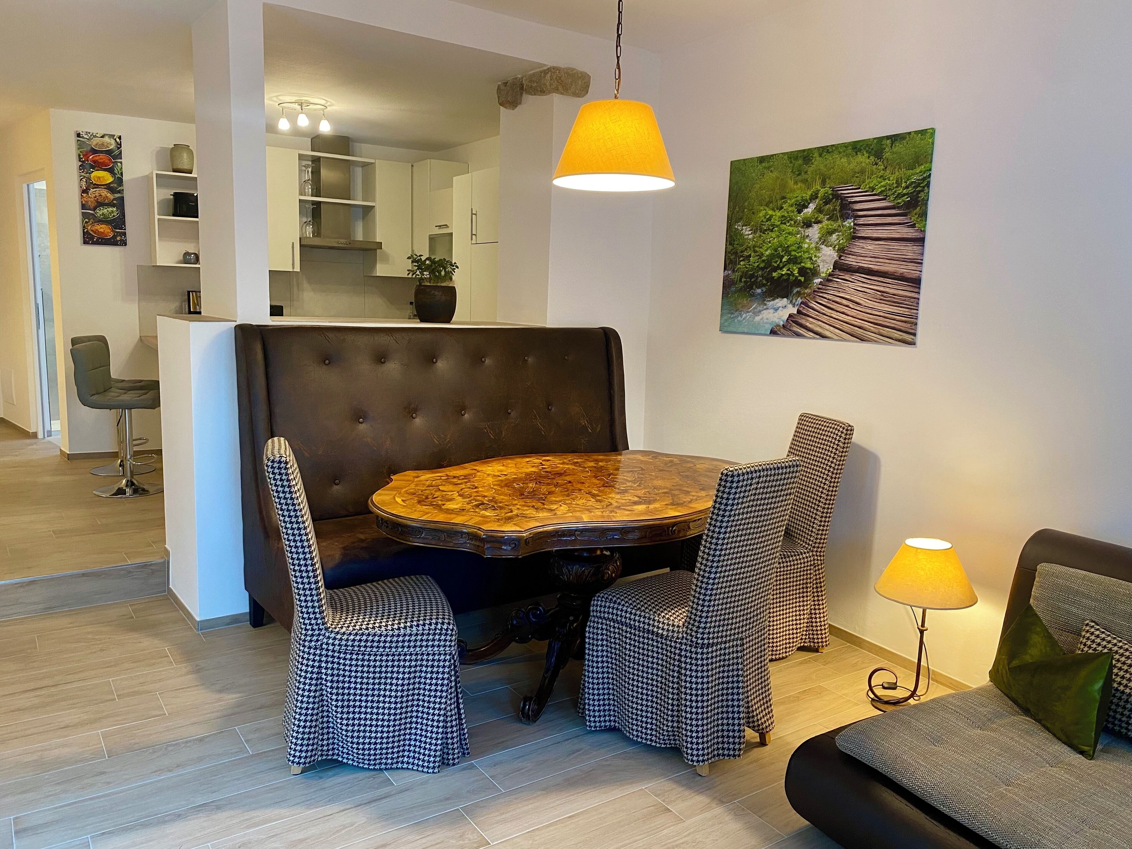Een eetgedeelte met een ronde tafel, stoelen met hondentanddessin, een bruine gestoffeerde bank en een gele hanglamp.