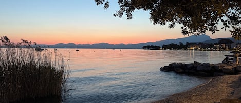 A calm lake at dusk with boats, a rocky shoreline, and distant mountains.