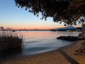 Un lago tranquilo al atardecer con barcos, una costa rocosa y montañas a lo lejos.