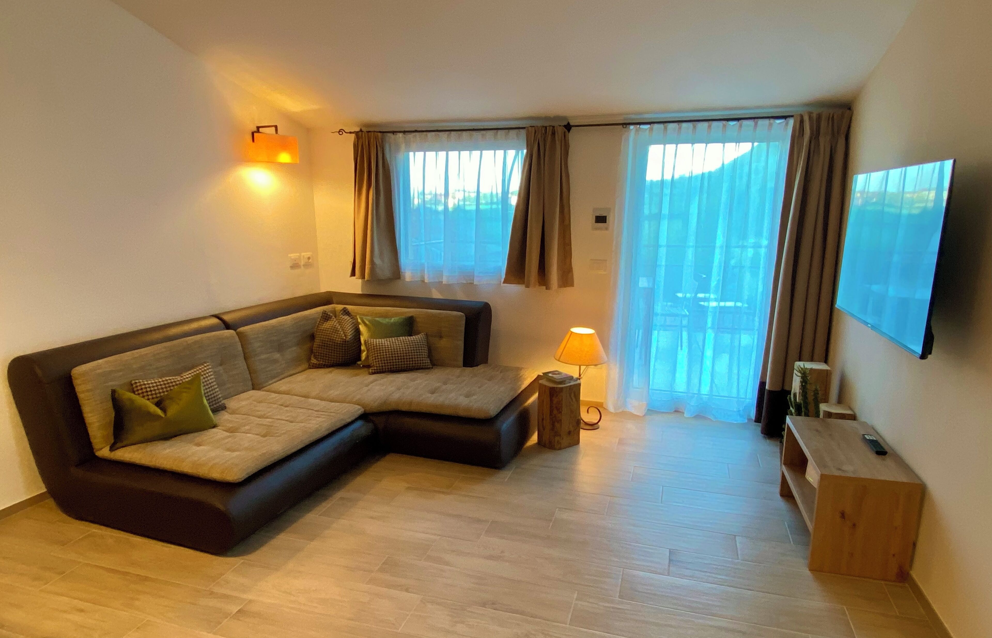 Uma sala de estar moderna com um sofá seccional, uma mesinha lateral, uma luminária, uma TV fixada na parede e uma grande janela com cortinas.