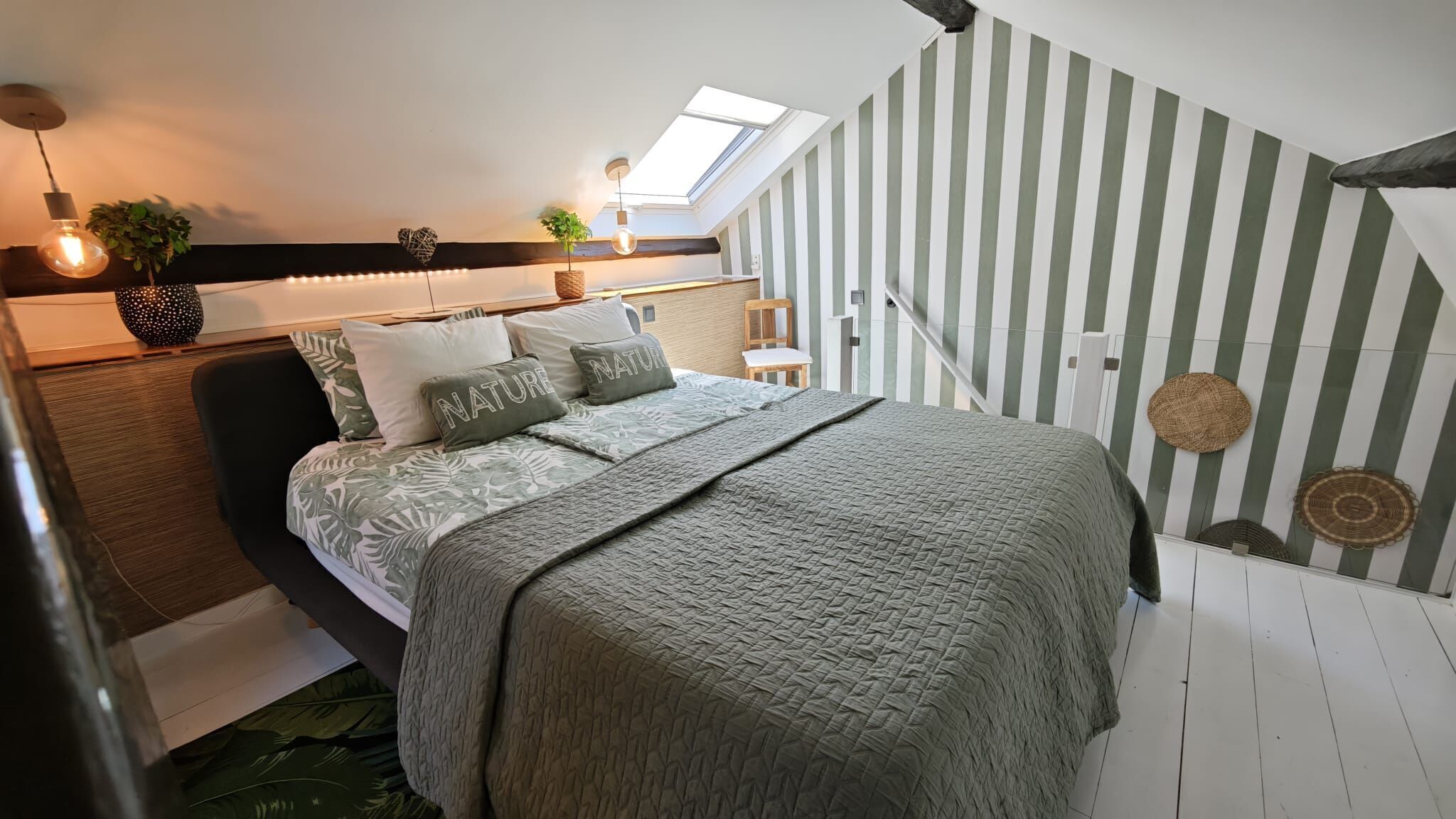 Une chambre avec un lit, une lucarne, un mur à rayures et une plante.