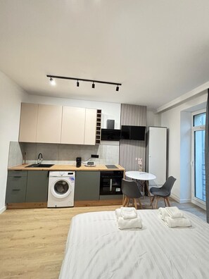 A compact, modern room with a bed, a small kitchen area including a washing machine, a dining table with chairs, and a large window providing natural light. - Apartamenty Styczyńskiego 2 (Katowice)