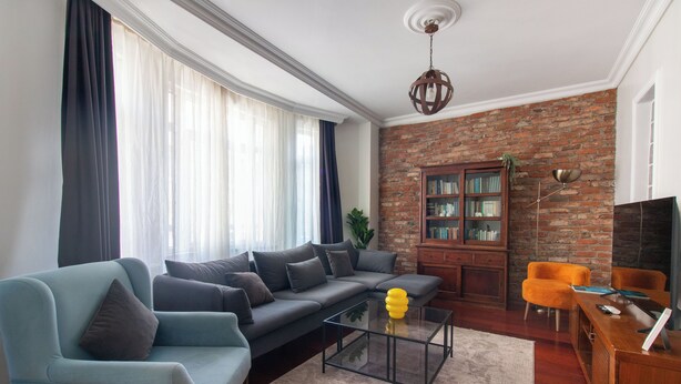 A living room with a sectional sofa, a glass coffee table, a bookshelf, and a brick wall. - Cozy 3BR Apartment in Şişli Close to Cevahir Mall (İstanbul)