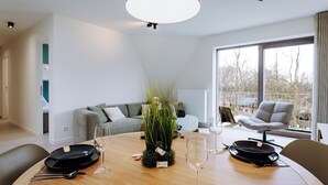 Un'area pranzo moderna con un tavolo rotondo in legno apparecchiata per quattro persone, un divano grigio e una grande finestra con vista sugli alberi.