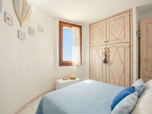 Ein Schlafzimmer mit Bett, Kleiderschrank, einem kleinen Tisch mit Hut und Sonnenbrille sowie einem Fenster mit Vorhängen.