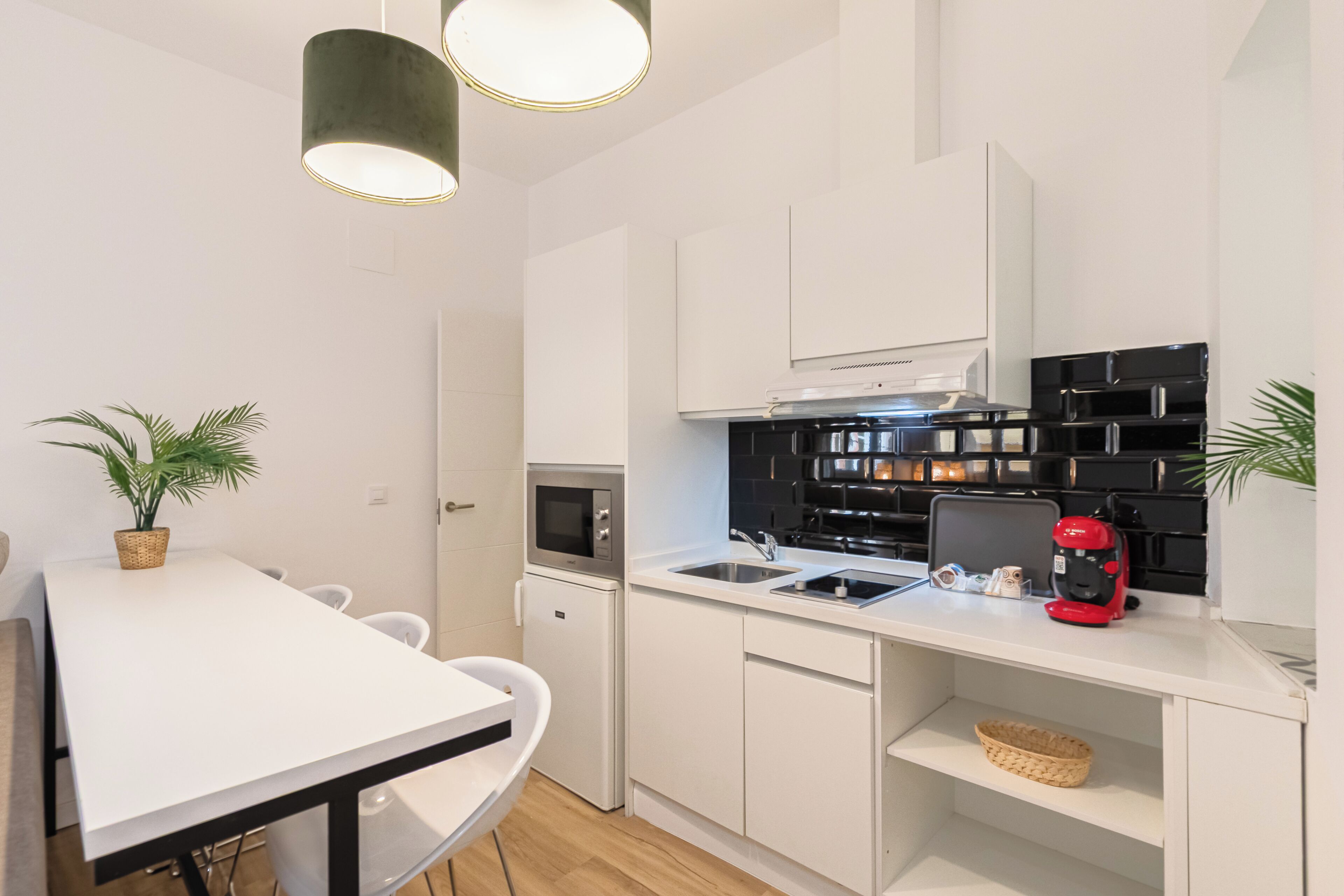 Una cocina moderna con armarios blancos, microondas, fregadero, cafetera y dos lámparas colgantes.