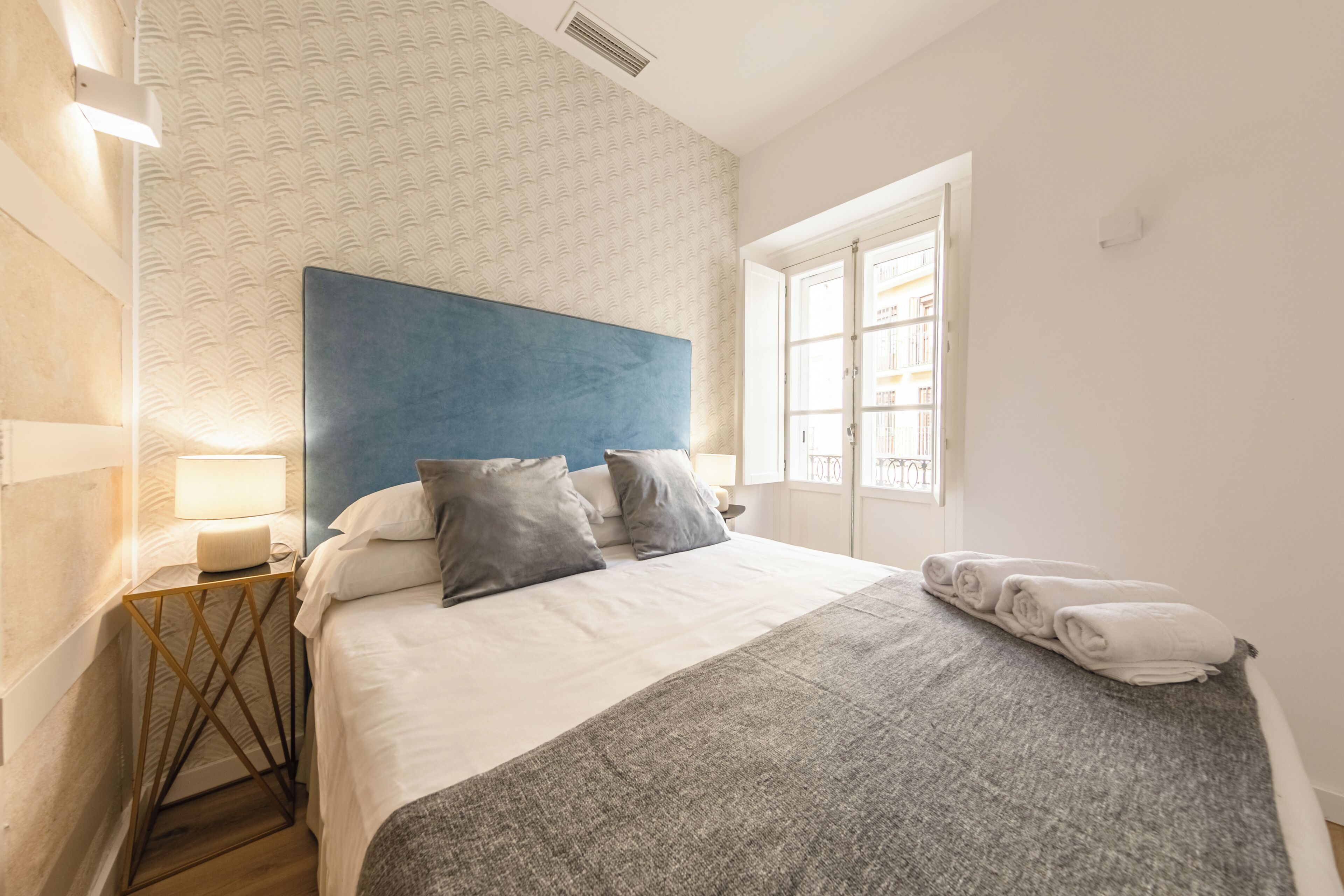 A neatly made bed with a blue headboard, a bedside lamp, and a window with white curtains.