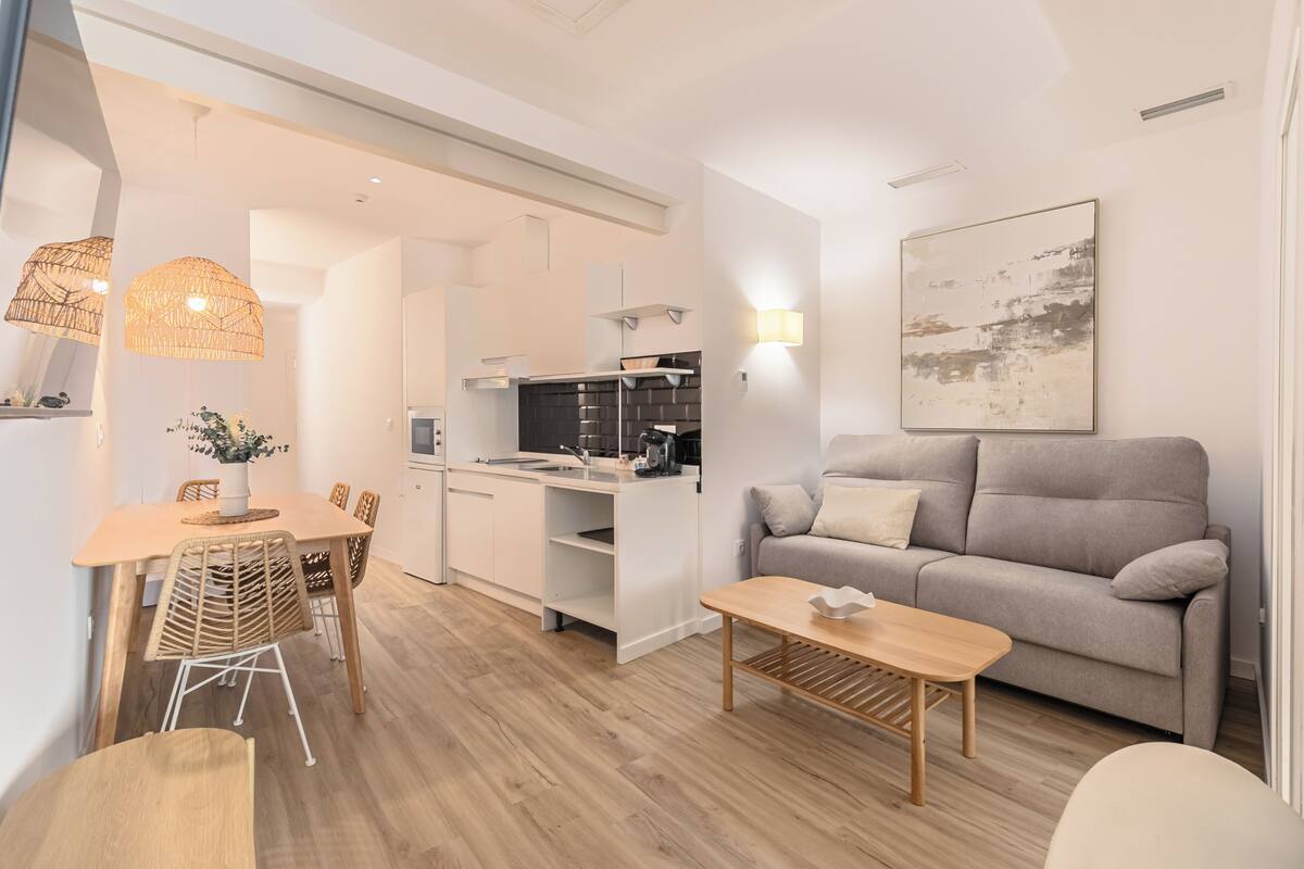 Una cocina moderna con armarios blancos, una zona de comedor con una mesa y sillas de madera, un sofá gris y una mesa de centro.
