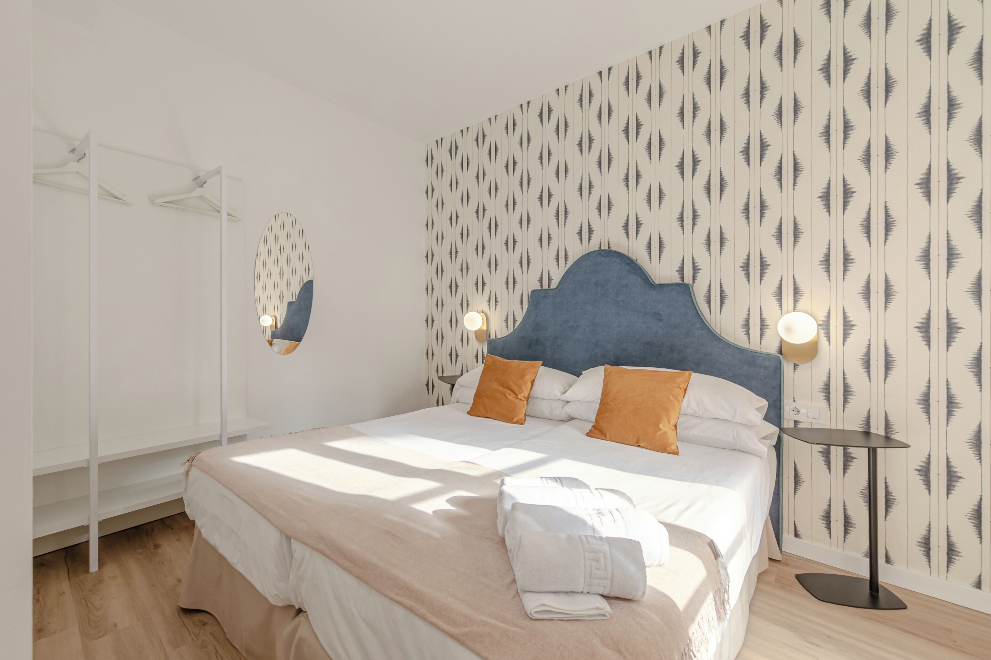A neatly made bed with a headboard, two pillows, a bedside table with a lamp, and a wall with a leaf pattern.