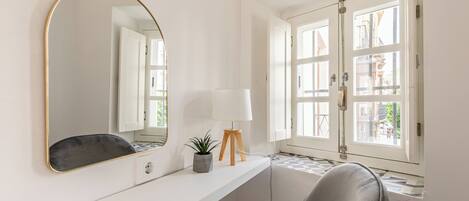 A bathroom with a large mirror, a small table with a lamp, a chair, and a window with a view of buildings.
