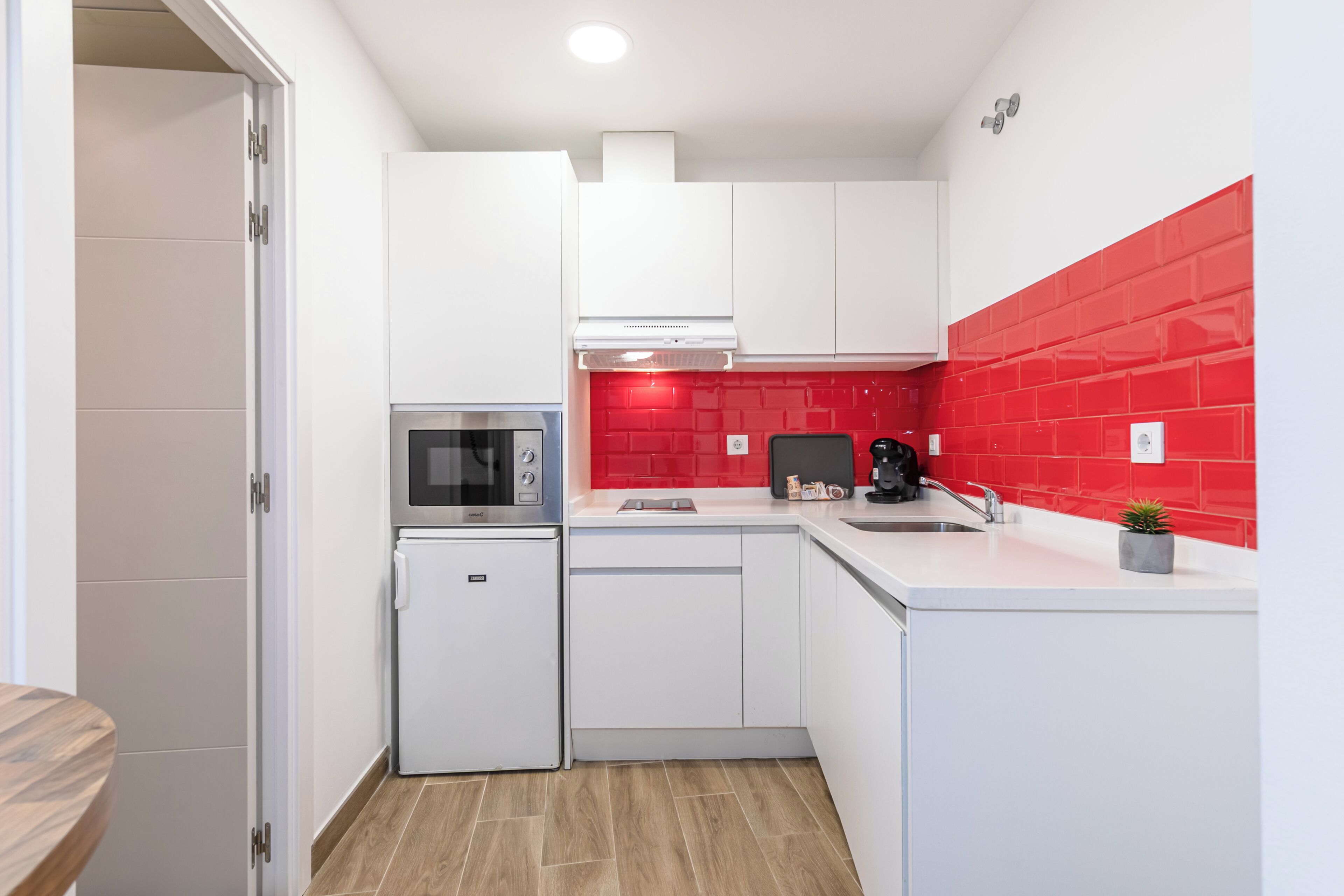Una cocina compacta con armarios blancos, un microondas, un refrigerador pequeño, un lavamanos y un salpicadero de azulejos rojos.