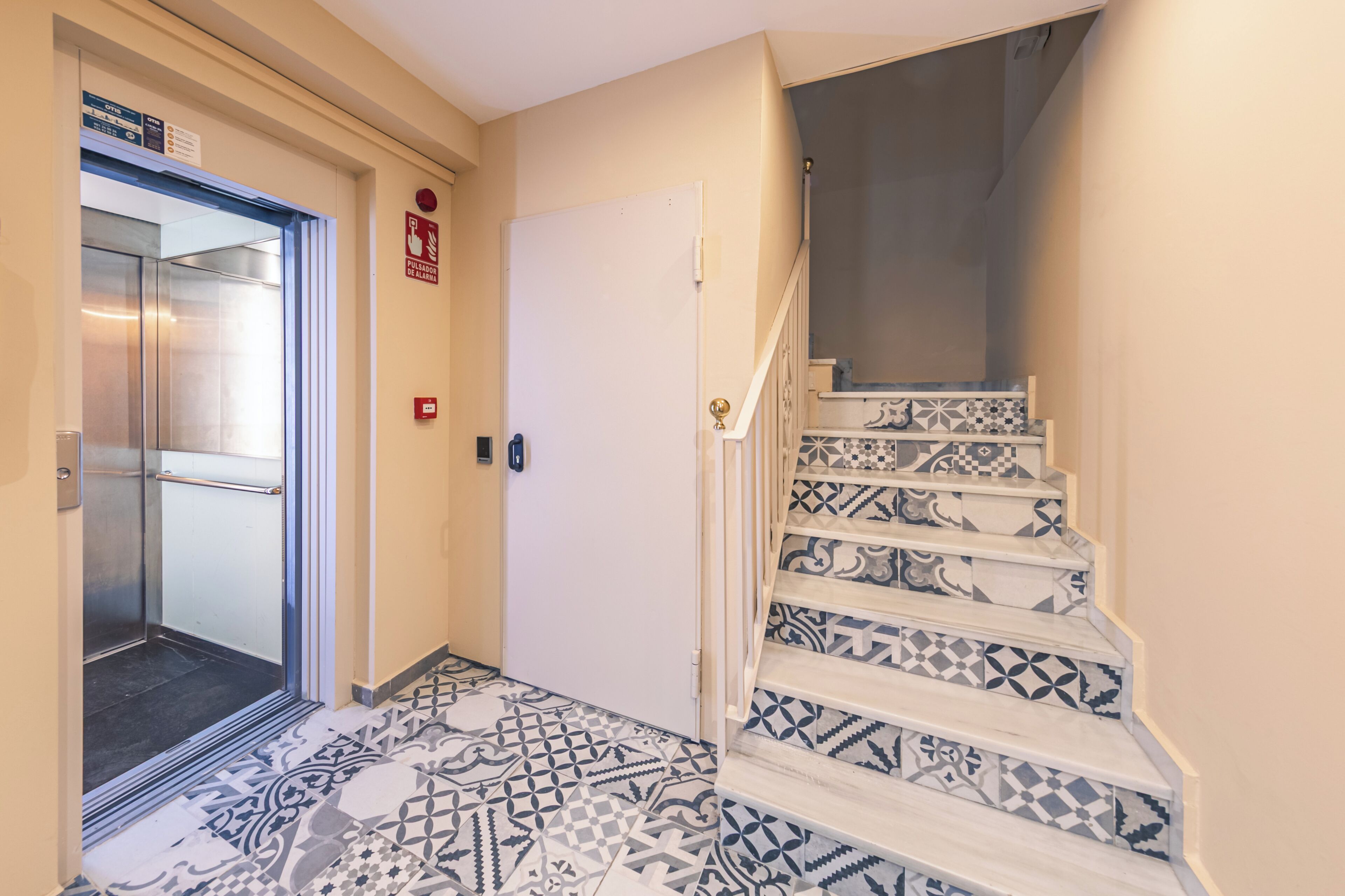 A hallway with a patterned floor, a door with a fire alarm sign, and a staircase with a patterned railing.
