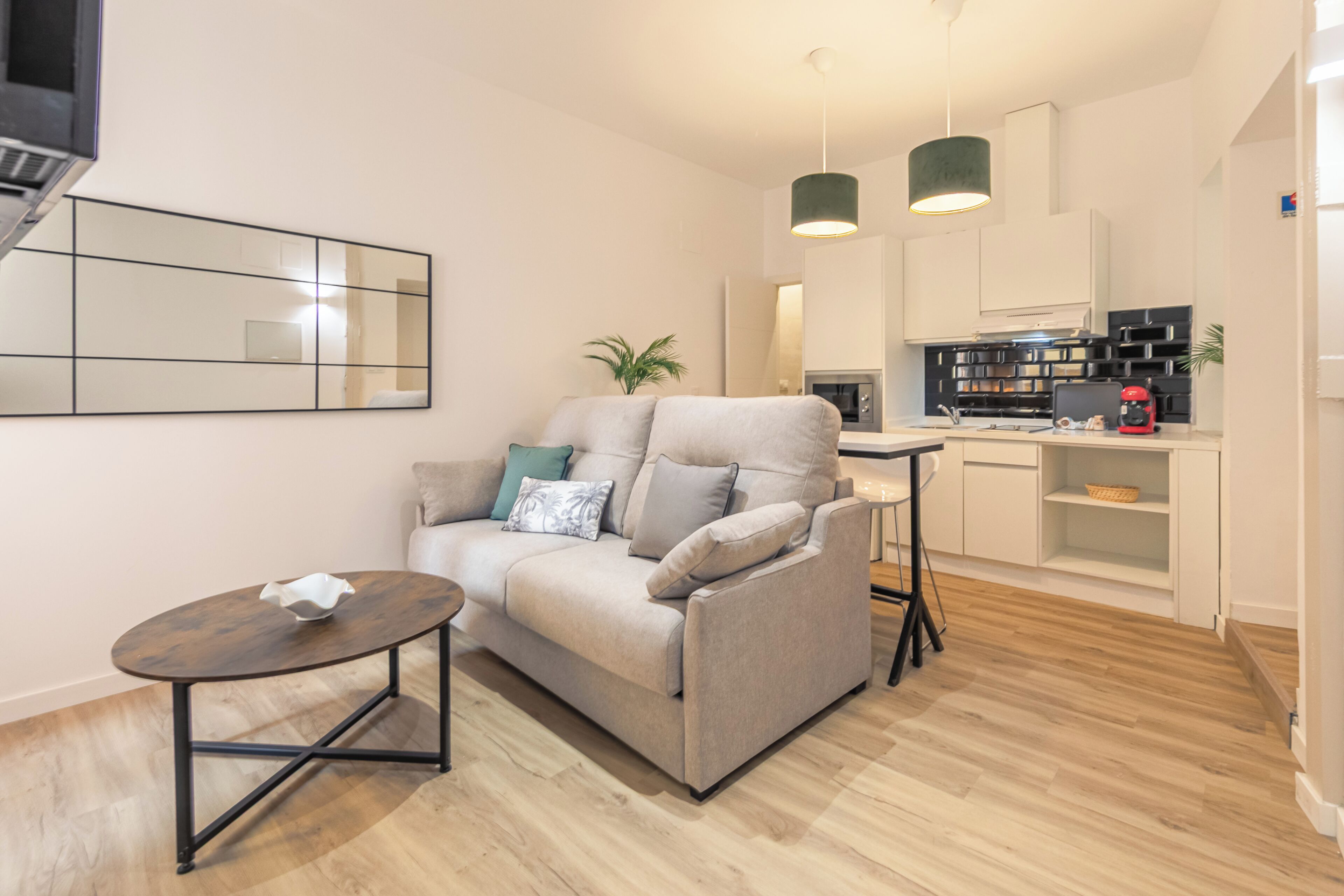 A modern living room with a sofa, a small round table, a kitchen area with cabinets and appliances, and a large mirror on the wall.
