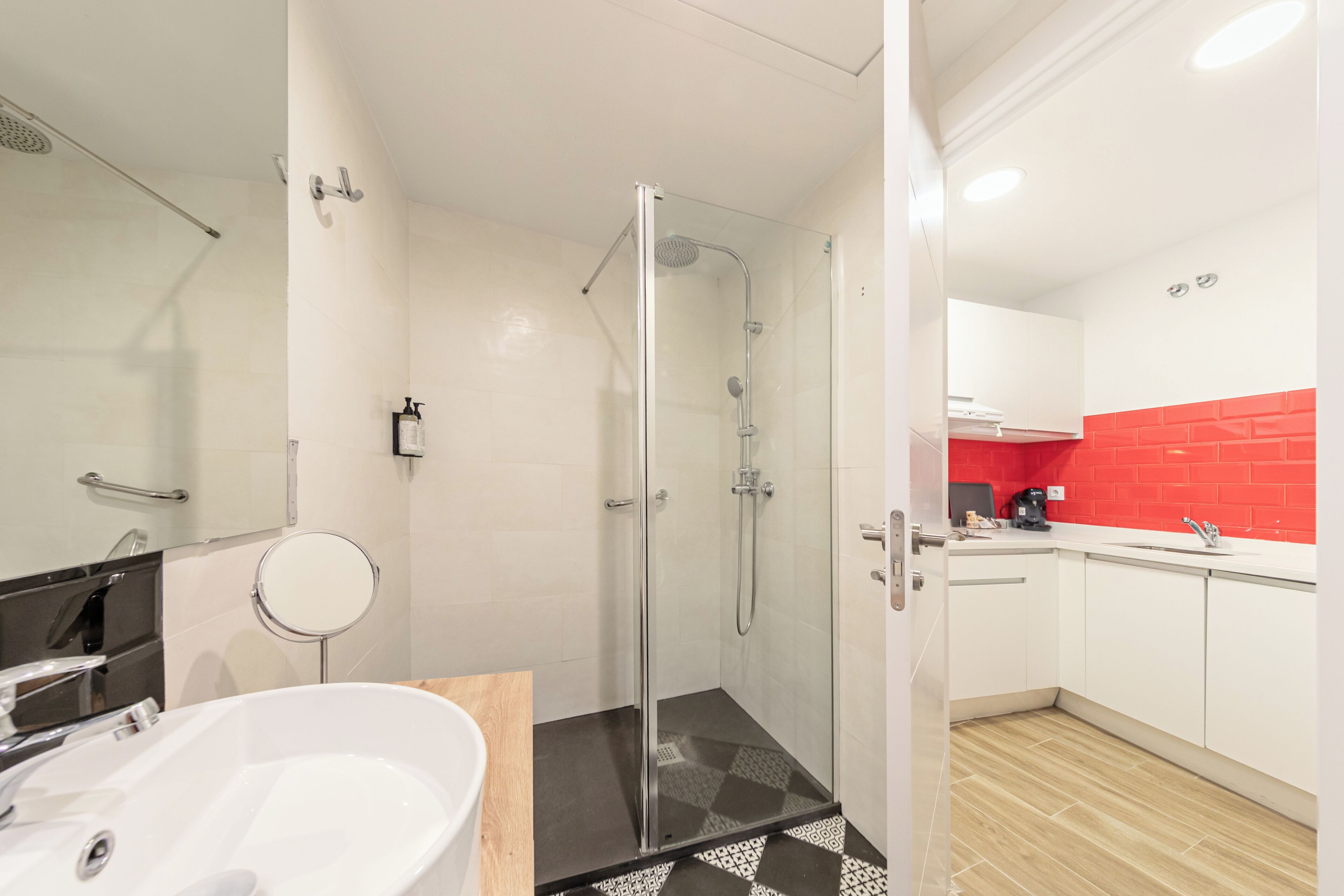 A modern bathroom with a glass-enclosed shower, a white sink, and a red-tiled backsplash in the adjacent kitchen area.