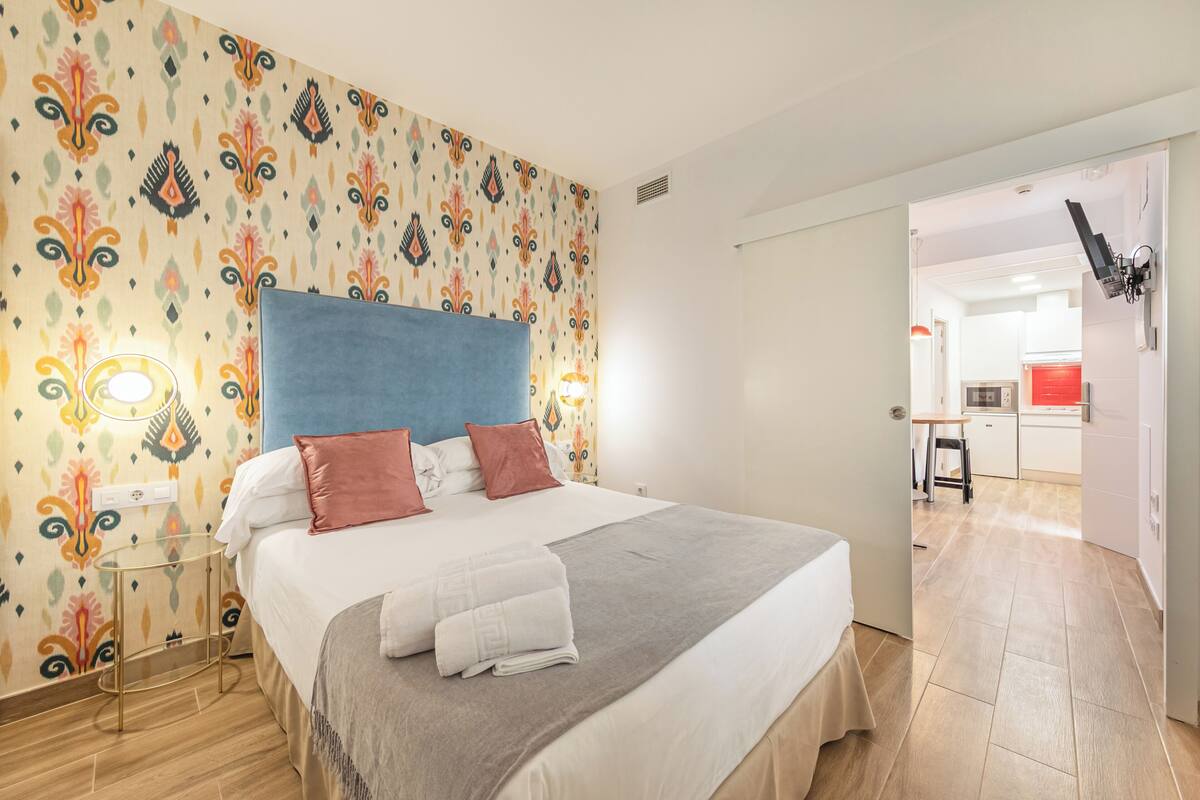 Habitación de hotel con cama, papel tapiz estampado, mesita de noche y vista a la cocina.