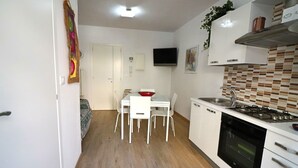 Una cucina compatta con mobili bianchi, un fornello e un lavandino. C'è un tavolo da pranzo con sedie e un televisore montato a parete.