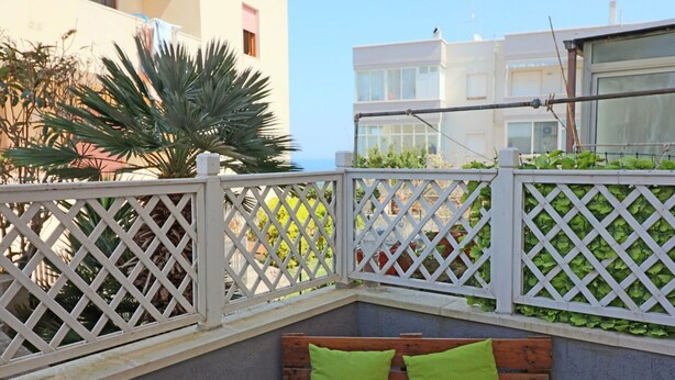 Un balcone angolare con un recinto in legno traforato, una panca in legno con cuscini verdi e vista su un edificio con balconi.