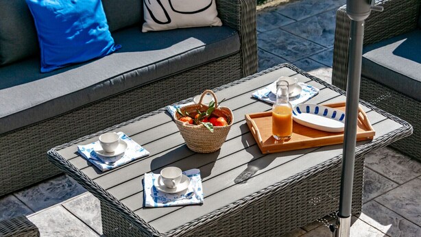 A wicker outdoor seating area with a table set for two, including a basket of fruit, a bottle of orange juice, and a tray with a cup and saucer. - Rental in Calpe REAL SITIO (Calpe)