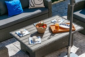 Un área de estar al aire libre con muebles de mimbre y una mesa preparada para dos, incluyendo una cesta de fruta, una botella de zumo de naranja y una bandeja con una taza y un plato.