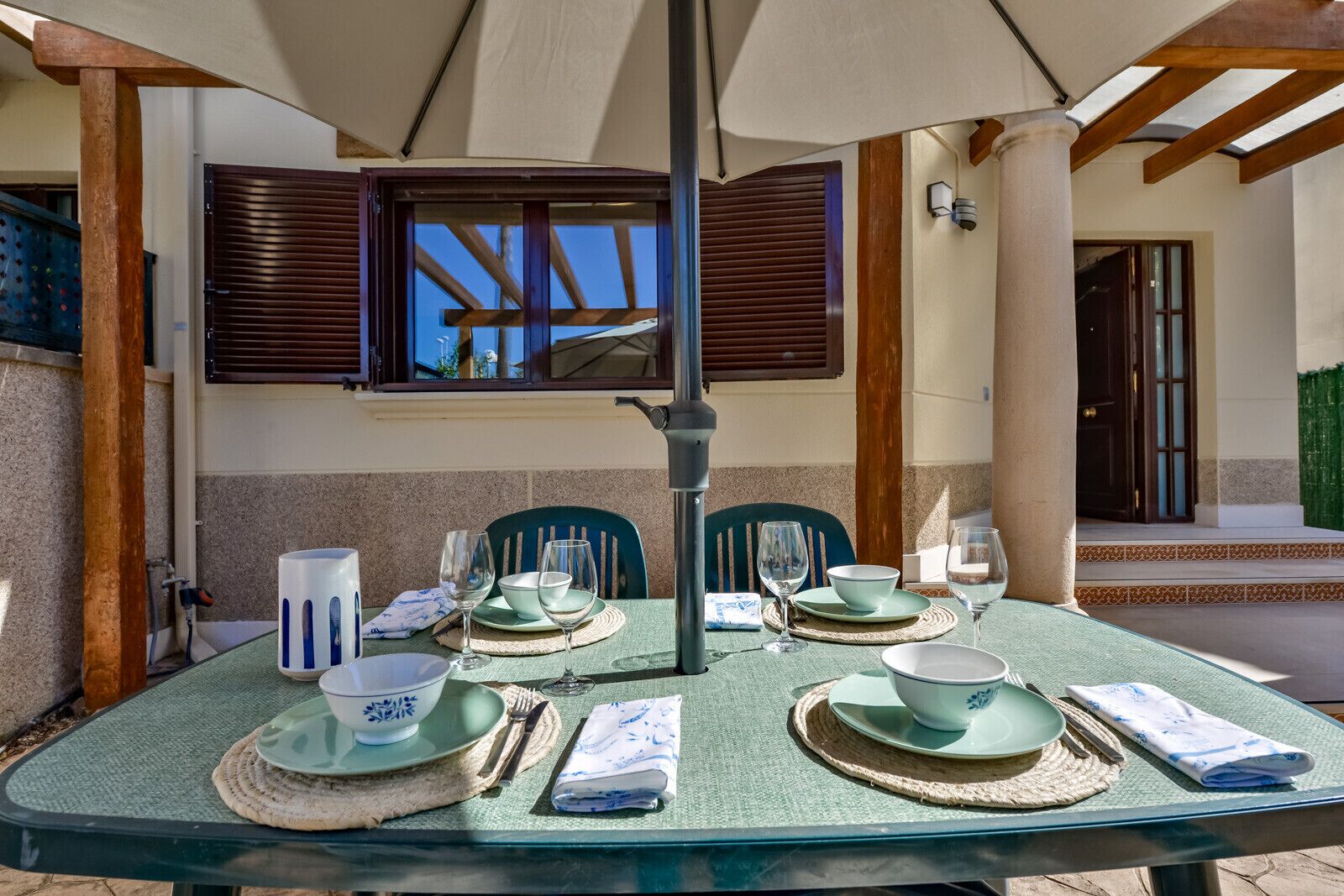 A dining table set outdoors with a table umbrella, chairs, and a view of the building's interior.