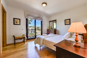 Une chambre d’hôtel avec un lit, un bureau avec une lampe, un banc, une commode et une grande fenêtre donnant sur l’extérieur.