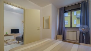 Une chambre à coucher avec un lit, une télévision, une fenêtre avec des rideaux, un radiateur et un ventilateur.
