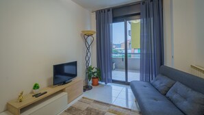 Un salon moderne doté d’un téléviseur à écran plat, d’un canapé, d’une petite table avec une plante et d’une vue sur un balcon extérieur, accessible par une porte-fenêtre coulissante.