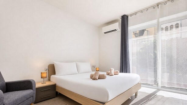 Ein modernes Hotelzimmer mit Bett, Nachttisch, Lampe und Blick auf die Stadt durch große Fenster.
