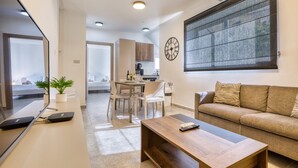 Ein modernes Wohnzimmer mit einer Couch, einem Couchtisch, einem Essbereich mit Stühlen, einer Küche im Hintergrund und einem großen Fenster mit Jalousien.