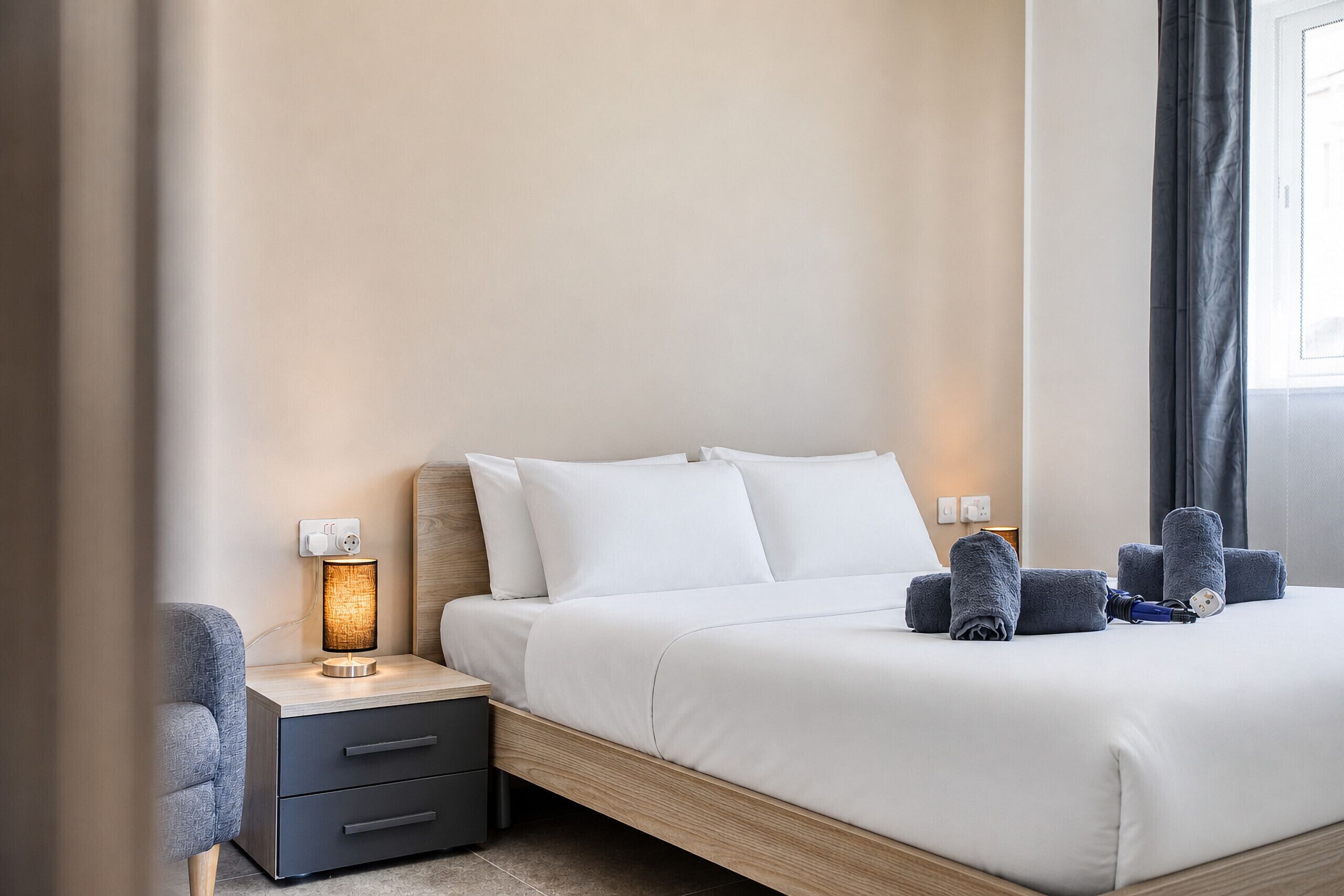 Een netjes opgemaakt bed met witte lakens, een bedlampje en een houten nachtkastje met een lade.