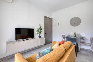 A modern living room with a flat-screen TV, a sofa, a dining table with chairs, and a clock on the wall. - Charming 2 bedroom Apartment, Valley Views KARP1-5 (San Gwann)