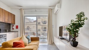 En moderne lejlighed med sofa, en stueplante, brandslukker og udsigt over byen gennem store vinduer.