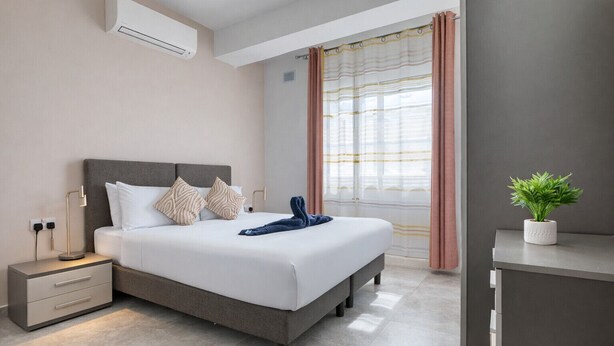 Una camera d'albergo moderna con un letto, comodino, lampada, condizionatore d'aria, finestra con tende e una piccola pianta.