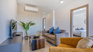 A modern living room with a flat-screen TV, a wooden coffee table, a sofa with blue cushions, a potted plant, and a bed in the background.