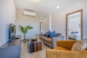 A modern living room with a flat-screen TV, a wooden coffee table, a sofa with blue cushions, a potted plant, and a bed in the background. - 2 bedroom flat with balcony, valley views KARP1-8 (San Gwann)