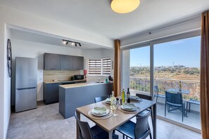 A modern kitchen with a central island, a dining area with a table set for two, and a balcony with a view of the city. - 2 bedroom flat with balcony, valley views KARP1-8 (San Gwann)