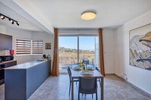 A modern kitchen with a dining area, a large window with curtains, and a view of the landscape. - 2 bedroom flat with balcony, valley views KARP1-8 (San Gwann)