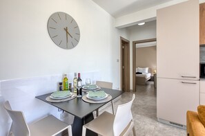 A modern dining area with a round wall clock, a table set for four, and a kitchen in the background. - Refined 2 bedroom apartment with balcony KARP1-9 (San Gwann)
