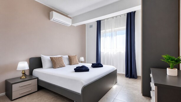 Ein modernes Hotelzimmer mit Bett, Nachttisch, Lampe, Klimaanlage, Vorhängen und einer kleinen Pflanze.