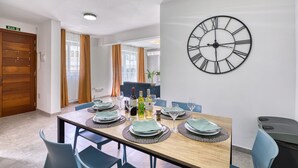 Un'area pranzo con un tavolo di legno apparecchiato per quattro persone, una grande orologio a parete e vista su un soggiorno attraverso una porta.