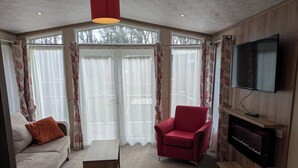 A room with a sofa, a red armchair, a wooden coffee table, a television mounted on a wooden wall, and large windows with sheer curtains.