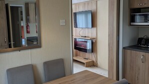 A compact living space with a wooden dining table, grey upholstered chairs, a flat-screen TV mounted on a wooden panel, a red pendant light, and a kitchenette with a microwave.