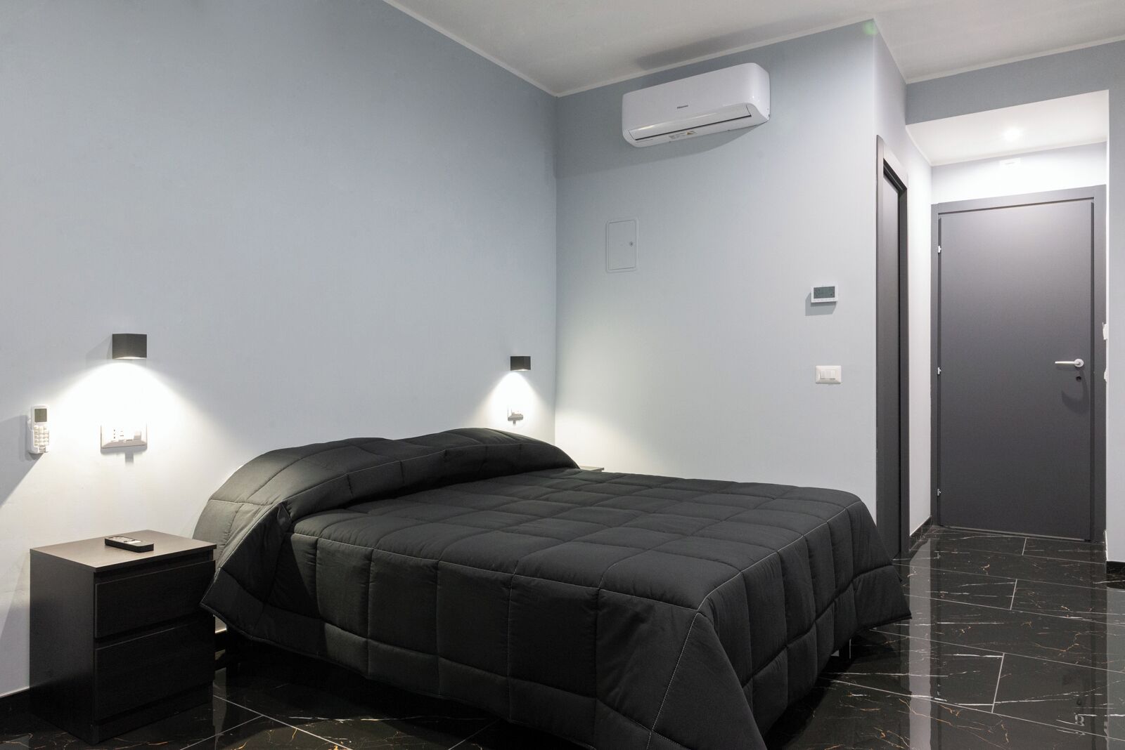 A modern bedroom with a black bed, a bedside table, a wall-mounted air conditioner, and a door.