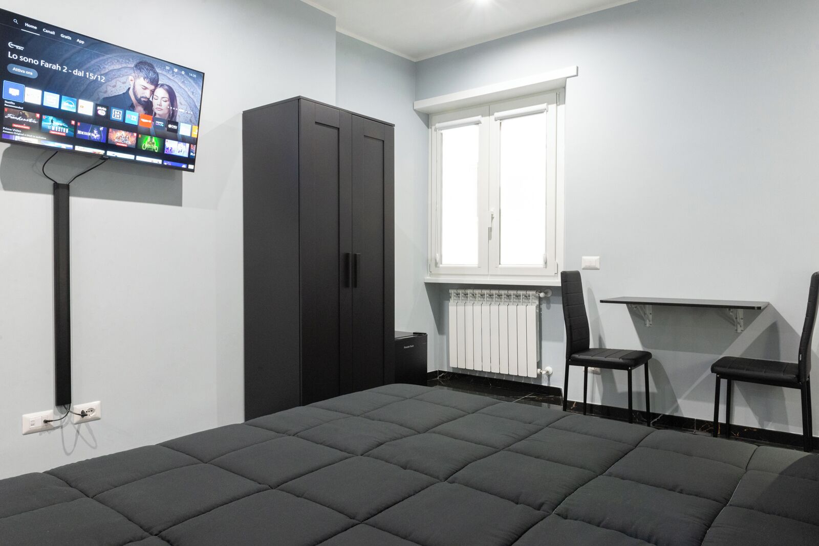 A modern room with a large bed, a TV mounted on the wall, a wardrobe, a radiator, and a small table with two chairs.