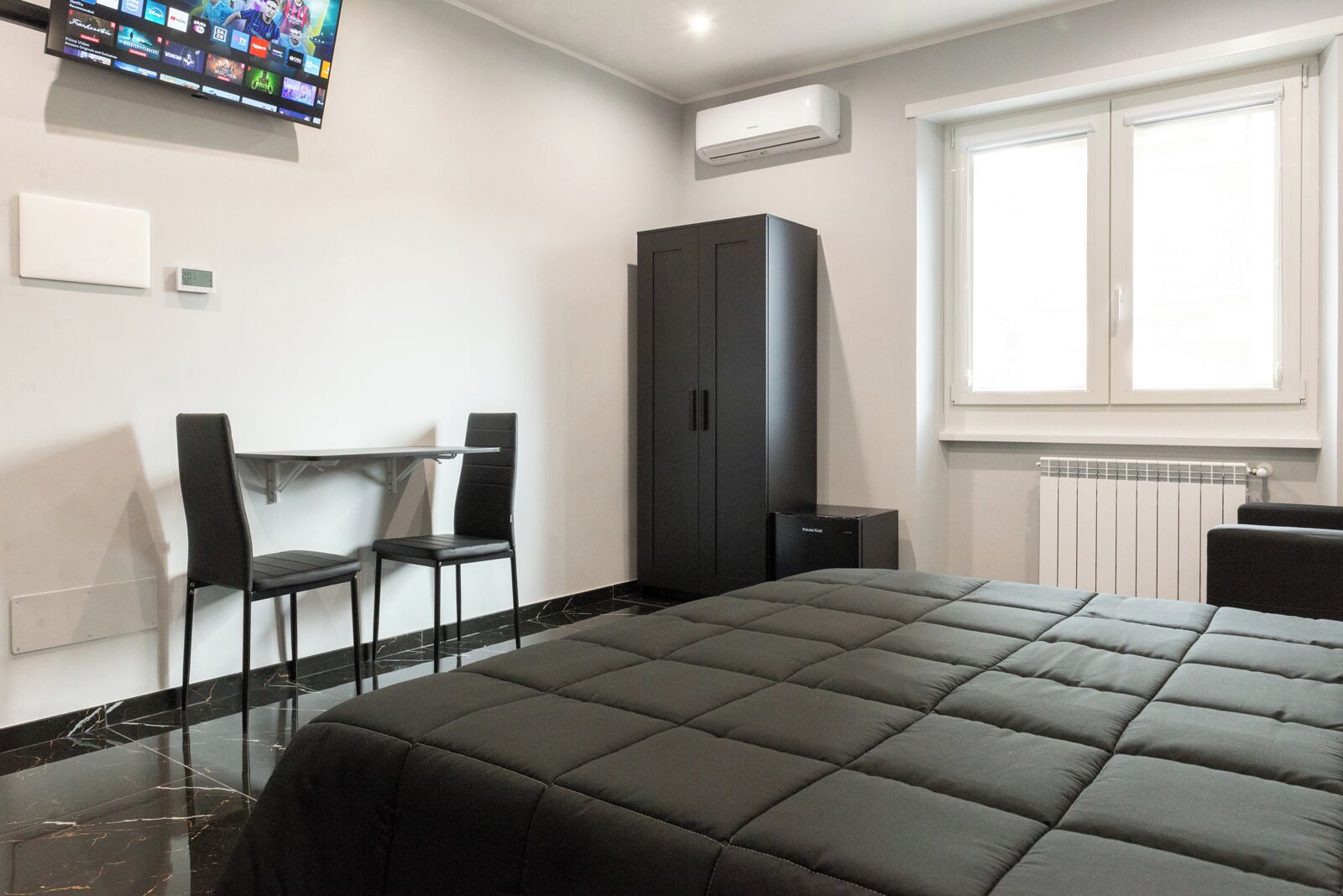 Un dormitorio moderno con una cama grande, un escritorio con dos sillas, un armario y un televisor montado en la pared.