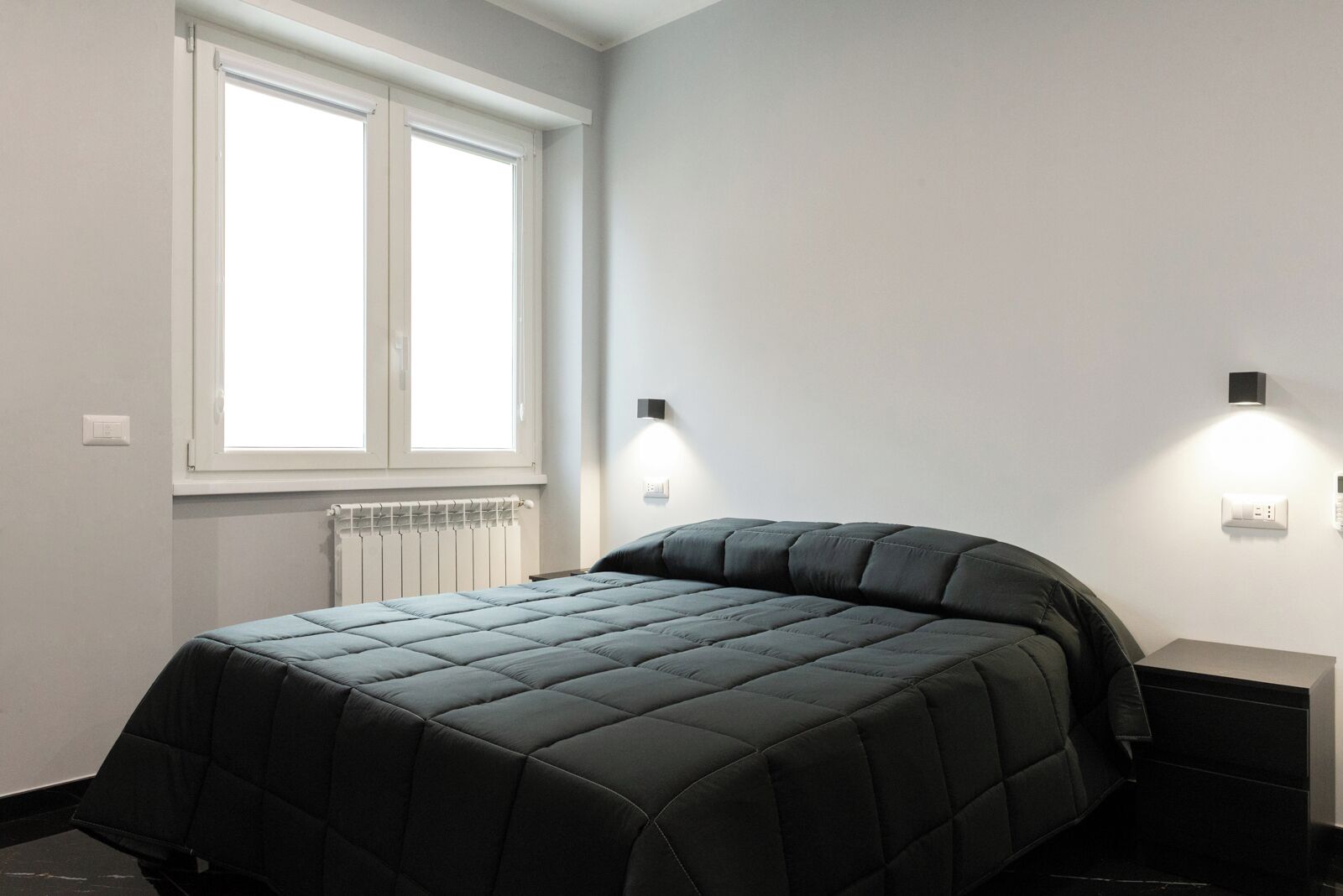 A bedroom with a bed, a window, a radiator, and two wall-mounted lights.