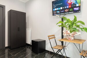 Una habitación moderna con una televisión de pantalla plana mostrando la interfaz de un servicio de streaming, un parlante negro, una mesa de madera con dos sillas, una planta en maceta y un radiador.