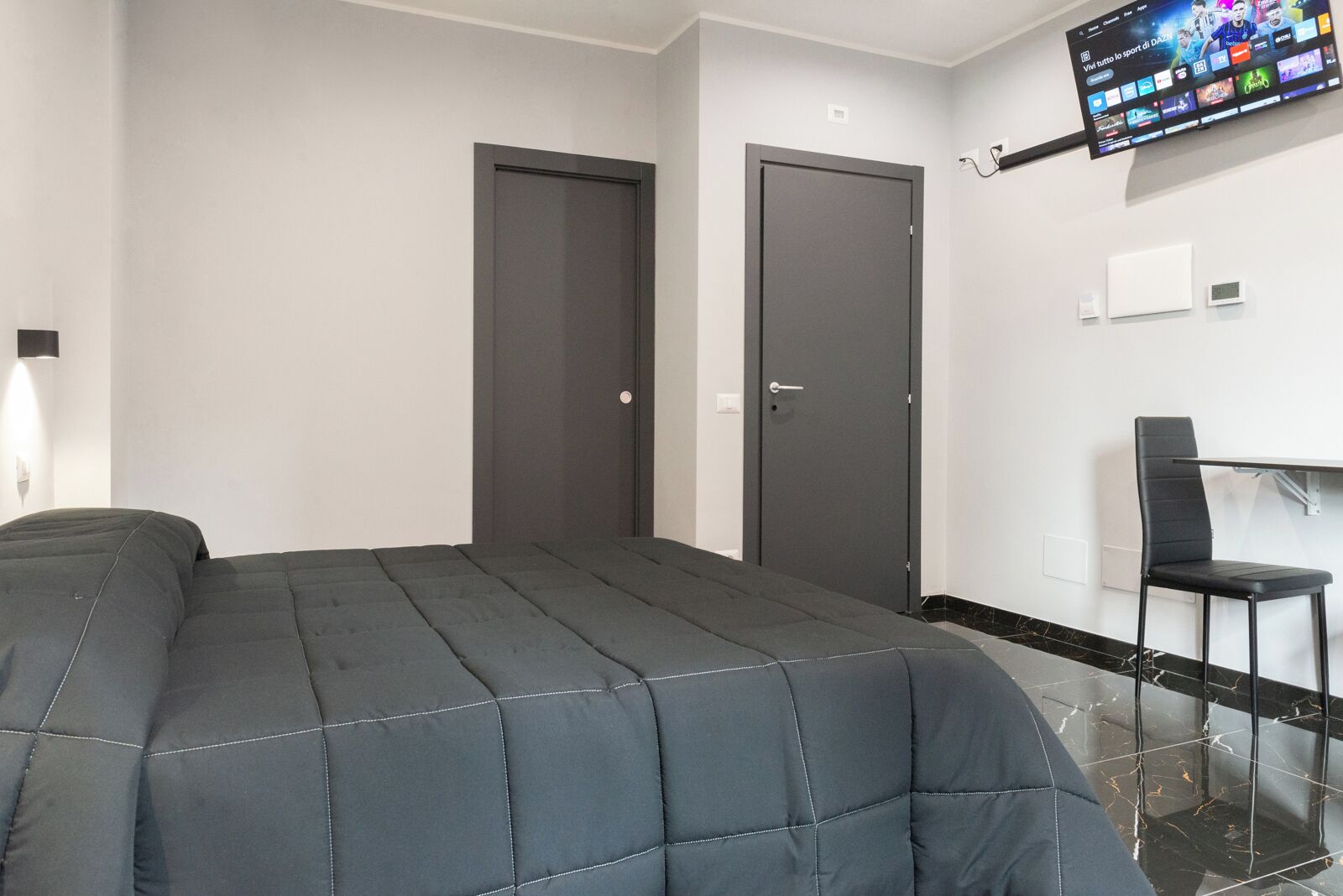 A modern hotel room with a bed, two doors, a TV mounted on the wall, and a chair.